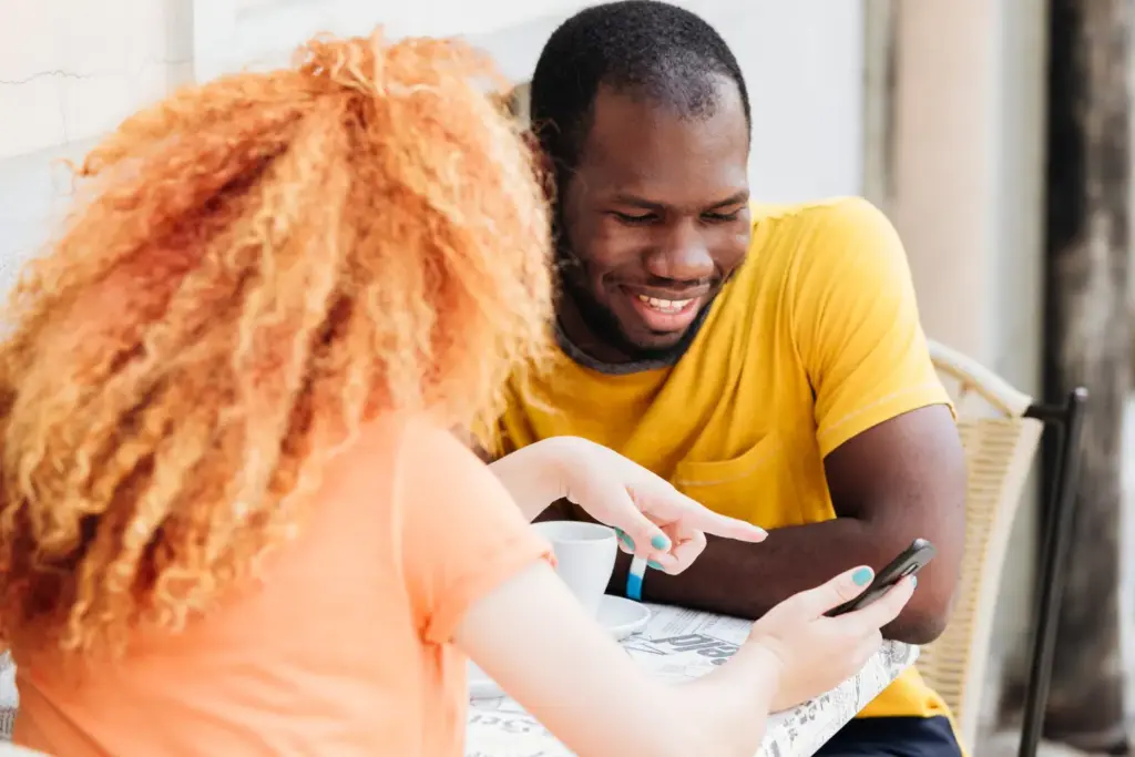 Smiling man and a woman with curly red hair sitting at a cafe table, looking at a smartphone screen to apply for Maine payday loans.