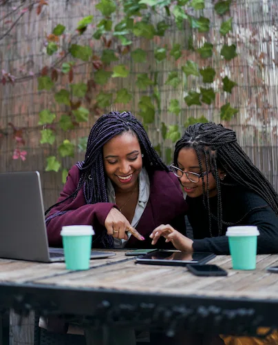 A woman in a burgundy jacket points at a tablet screen while discussing payday loans in Maryland with a friend using a laptop at an outdoor table