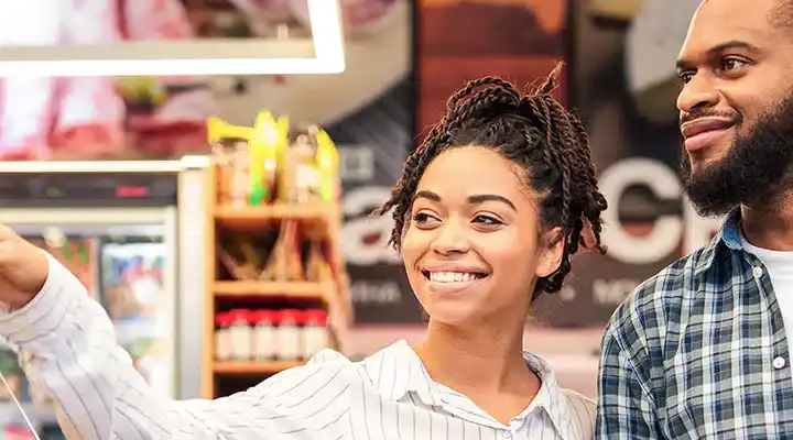 A woman with curly hair in a white top points towards colorful vegetables on a display in a grocery store. Next to her, a man in a black and white checkered shirt stands, looking at the display with interest.