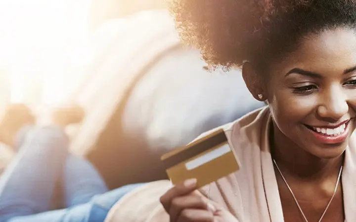 A woman with curly hair lying on a couch holds a credit card with her left hand. The background features a softly lit living area with a blurred view of furniture.